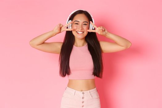 Portrait of beautiful smiling asian girl in wireless headphones, listening music, enjoying sound quality, showing peace or kawaii gestures voer eyes and grinning happy over pink background.