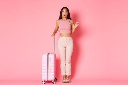 Travelling, holidays and vacation concept. Silly and cute asian girl in summer clothes meeting girlfriends in airport, packed bags for paradise resort journey, holding suitcase over pink background.
