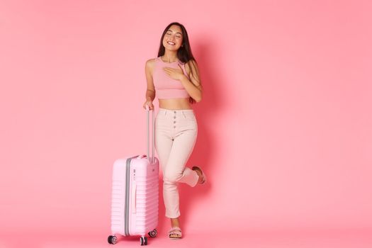 Travelling, holidays and vacation concept. Full-length of dreamy, silly asian girl daydreamign about future travel, tourist dreaming about paradise resort, standing with suitcase, pink background.