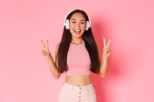 Portrait of beautiful and upbeat, smiling asian girl listening music in headphones, showing peace kawaii gesture, sending positive vibes, standing over pink background.