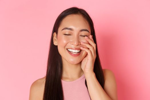 Beauty, fashion and lifestyle concept. Close-up of carefree beautiful asian girl touching face and laughing happily with closed eyes, standing over pink background joyful, skincare product promo.