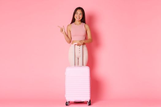 Travelling, holidays and vacation concept. Full-length of pleased attractive asian girl tourist in summer outfit, pointing finger left and looking with smile, stand with suitcase.