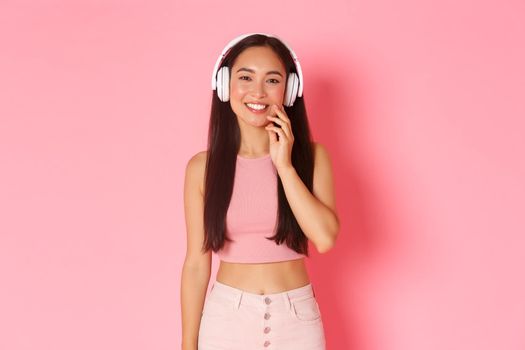 Portrait of upbeat, smiling pretty asian girl looking motivated with interesting podcast in headphones. Woman listening music and being pleased, standing over pink background.