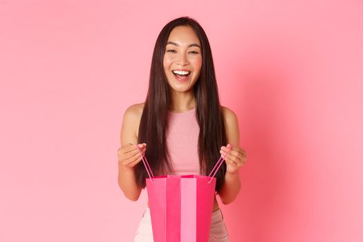 Beauty, fashion and lifestyle concept. Gorgeous happy asian girl receive gift, holding shopping bag and smiling dreamy, looking thankful and curious what inside, standing over pink background.