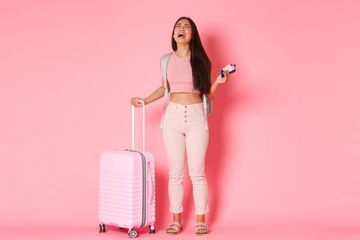 Travelling, holidays and vacation concept. Full-length of crying, sad and distressed asian girl tourist, feeling depressed about missed flight, standing suitcase and plane tickets, pink background.