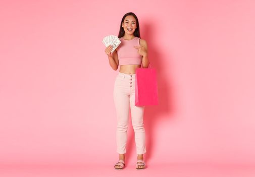 Full length of rich attractive asian girl in summer clothes, enjoying shopping, have money, pointing finger at cash to waste on shops and stores, standing pink background.