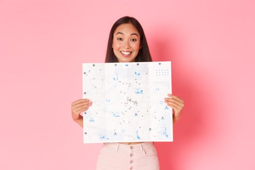 Travelling, lifestyle and tourism concept. Cheerful, attractive asian girl tourist explore new city, visiting museums, showing map of city with sightseeings and smiling upbeat, pink background.