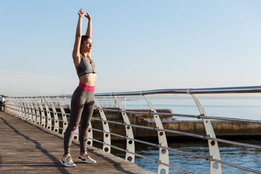 Young fit sportswoman stretching before morning exercises. Female runner workout on seaside promenade.