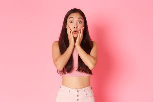 Lifestyle, beauty and women concept. Portrait of intrigued and surprised asian pretty girl see awesome discount offer, holding hands on cheeks and pouting excited, saying wow, pink background.
