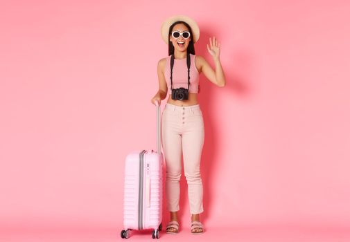 Tourism, summer vacation, holidays abroad concept. Full length of beautiful relaxed asian girl tourist, traveller in sunglasses and summer clothes waving hand, saying hello, pink background.