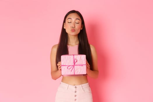Holidays, celebration and lifestyle concept. Lovely and silly pretty asian girl receiving wrapped gift, close eyes and kissing person who gave it to her, standing over pink background.