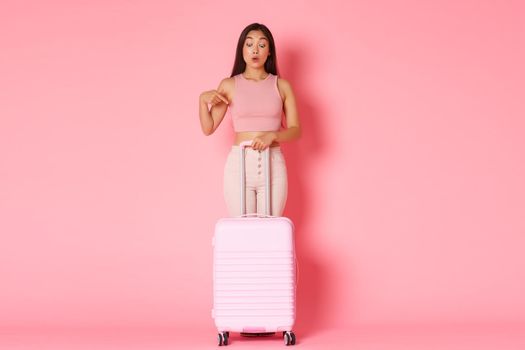 Travelling, holidays and vacation concept. Full-length of curious and interested asian tourist girl in summer clothes, holding suitcase, pointing finger down wondered, pink background.