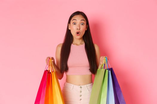 Beauty, fashion and lifestyle concept. Close-up of amused and excited pretty asian girl with shopping bags, impressed with lots of discounts and sales in stores, pink background.