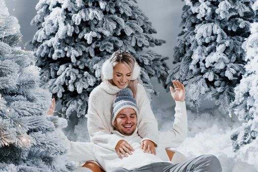 New year love story. Couple kisses and hugs. People weared wearing fur headphones, hats, white sweaters. Happy young couple hugs and kiss near christmas trees in winter day.