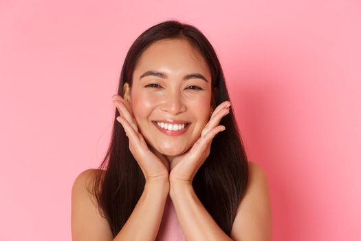 Beauty, fashion and lifestyle concept. Close-up of beautiful asian girl touching her face and smiling silly, blushing, feeling refreshed, apply skincare product or makeup, pink background.