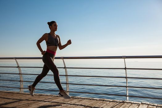 Side view of young fitness woman running along pier. Female athlete workout and jogging on the seaside promenade.