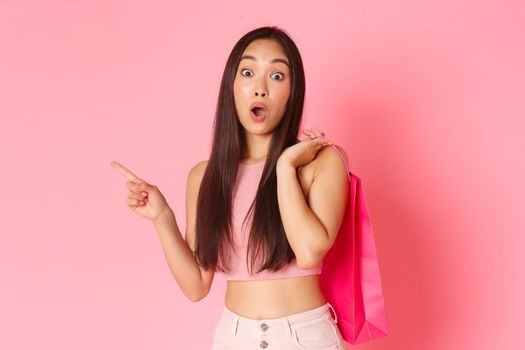 Beauty, fashion and lifestyle concept. Portrait of trendy asian girl enjoying shopping, holding bag over shoulder and pointing finger left with impressed, curious expression, want to check out.
