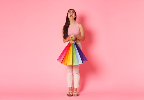 Full length portrait of crying and disappointed, whining asian girl complaining about not enough money, holding shopping bags and sobbing upset, standing pink background.