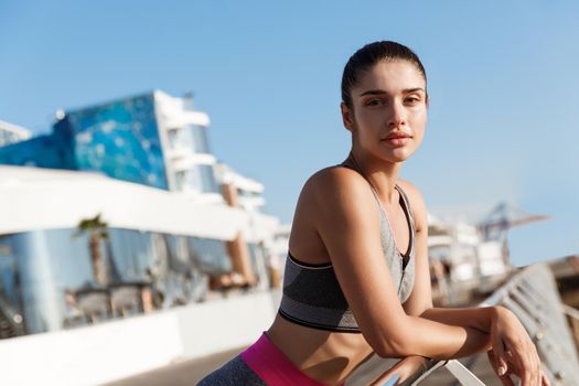 Close-up of attractive female runner standing on a pier with pleased face, workout outdoors near sea.