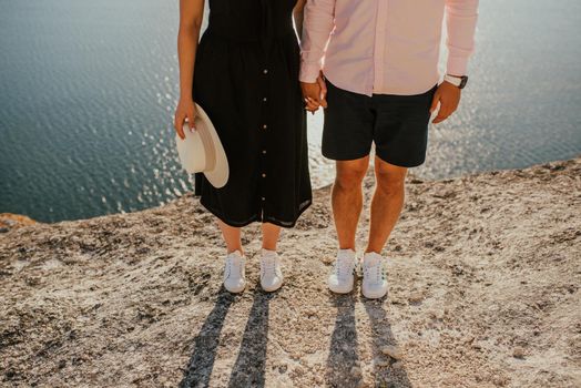 Male and female legs in white sneakers on a rock against the background of the sea. A couple of fair-haired fair-skinned people in love are resting in nature in a field at sunset.