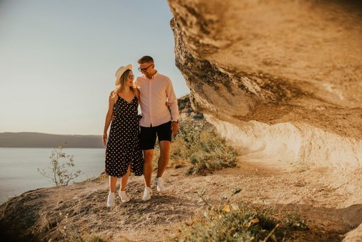 A man and a woman in a hat walk along the rocks above the cliff. A couple of fair-haired fair-skinned people in love are resting in nature in a field at sunset.