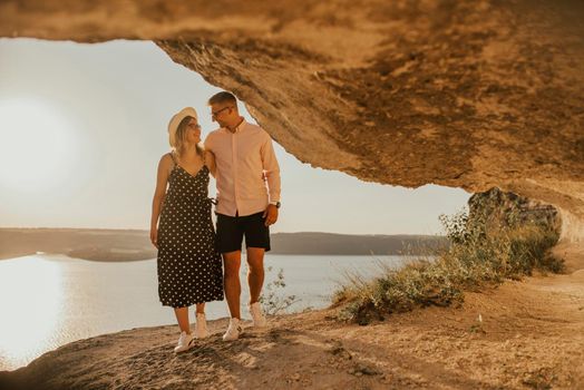 A man and a woman in a hat walk along the rocks above the cliff. A couple of fair-haired fair-skinned people in love are resting in nature in a field at sunset.