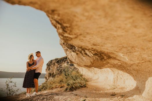 A man and a woman in a hat walk along the rocks above the cliff. A couple of fair-haired fair-skinned people in love are resting in nature in a field at sunset.