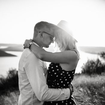 A man with a woman in a hat hug and kiss in the tall grass in the meadow. A couple of fair-haired fair-skinned people in love are resting in nature in a field at sunset.