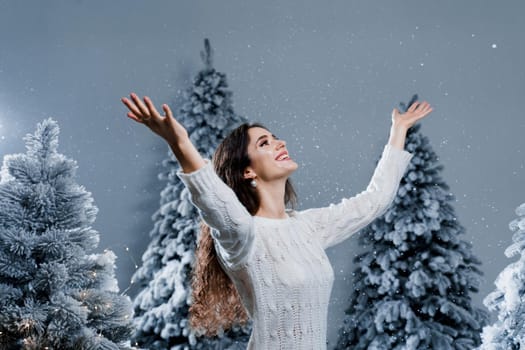 Happy girl smiles and touches falling snow at eve new year. Young woman near big snowy spruces in winter day before christmas celebration