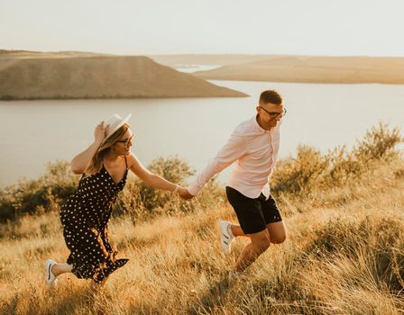 young man and woman walking in the meadow at sunset in summer near the lake. A couple of fair-haired fair-skinned people in love are resting in nature in a field at sunset.