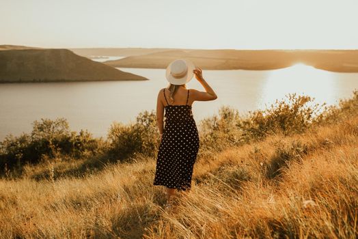 young woman in hat in dress with polka dots stands in middle of meadow on cliff above the sea at sunset