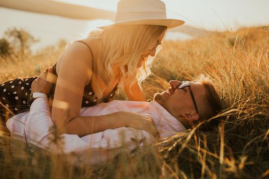 A man and a woman in a hat lie on the ground in the middle of the grass. A couple of fair-haired fair-skinned people in love are resting in nature in a field at sunset.