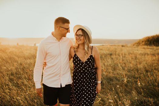 a guy with a girl in hat walking in the meadow. A couple of fair-haired fair-skinned people in love are resting in nature in a field at sunset.