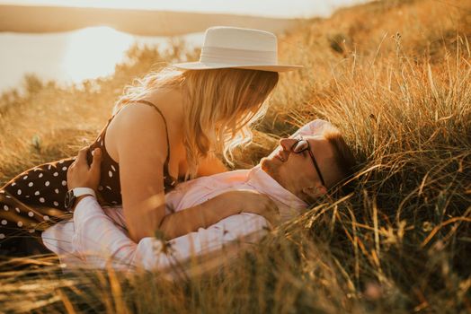 A man and a woman in a hat lie on the ground in the middle of the grass. A couple of fair-haired fair-skinned people in love are resting in nature in a field at sunset.