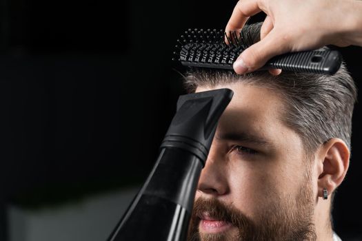 Barber dries the hair of a handsome bearded man after a fashionable haircut. The work of a hairdresser during the quarantine period coronavirus covid-19