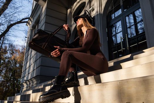 Fashionable young brunette woman with long legs. wearing solid long sleeve bodycon one piece jumpsuits posing with a leather brown travel bag on city street in old town
