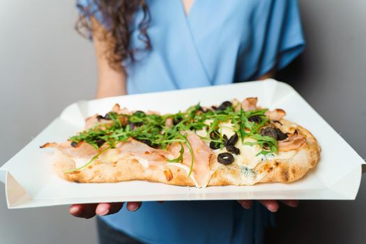 Pinsa romana in white delivery box. Courier holding crocchiarella gourmet italian cuisine on white background. Food delivery from pizzeria. Pinsa with meat, arugula, olives, cheese