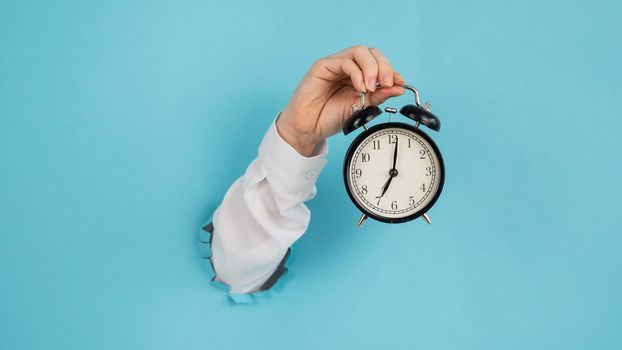 A woman's hand sticks out of a hole in a blue paper background and holds an alarm clock