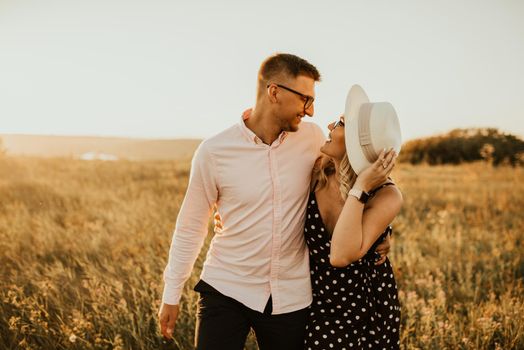 a guy with a girl in hat walking in the meadow. A couple of fair-haired fair-skinned people in love are resting in nature in a field at sunset.