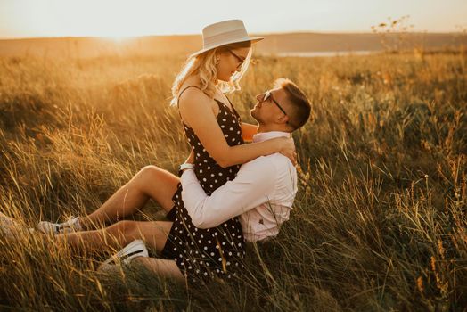 A girl in hat sits in hands guy on ground in middle of tall grass in meadow. couple of fair-haired fair-skinned people in love are resting in nature in field at sunset.