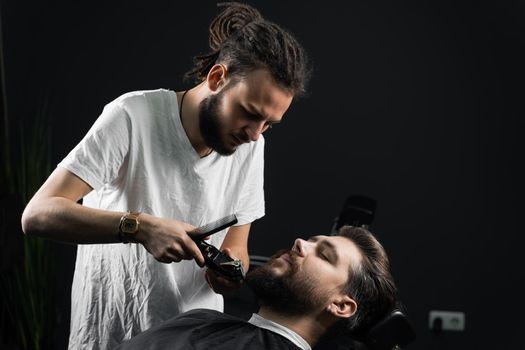 Beard shaving in barbershop. Barber with dreadlocks trim handsome bearded man