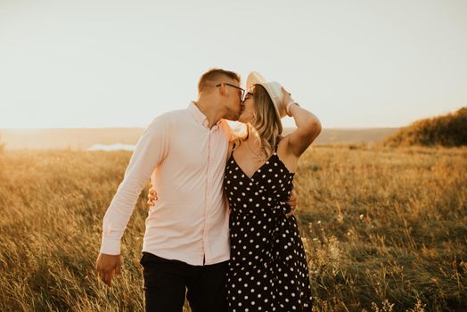 a guy with a girl in hat walking in the meadow. A couple of fair-haired fair-skinned people in love are resting in nature in a field at sunset.