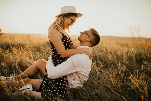 A girl in hat sits in hands guy on ground in middle of tall grass in meadow. couple of fair-haired fair-skinned people in love are resting in nature in field at sunset.