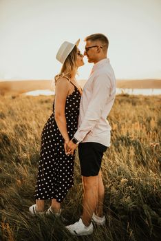 A man with a woman in a hat hug and kiss in the tall grass in the meadow. A couple of fair-haired fair-skinned people in love are resting in nature in a field at sunset.