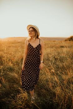 A fair-skinned blonde girl in a dress with polka dots in a hat walks through the meadow among the tall grass in the summer at sunset.