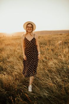 A fair-skinned blonde girl in a dress with polka dots in a hat walks through the meadow among the tall grass in the summer at sunset.