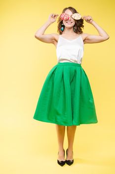 Full length studio portrait of laughing model in white top and green skirt and heels holding two sweet candies on her eyes like sunglasses with her mouth open. Isolate on yellow.