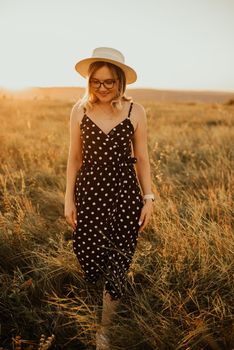 A fair-skinned blonde girl in a dress with polka dots in a hat walks through the meadow among the tall grass in the summer at sunset.