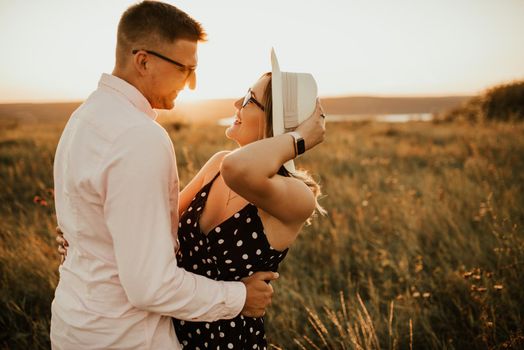 A man with a woman in a hat hug and kiss in the tall grass in the meadow. A couple of fair-haired fair-skinned people in love are resting in nature in a field at sunset.