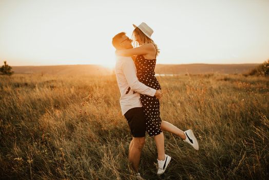 A man with a woman in a hat hug and kiss in the tall grass in the meadow. A couple of fair-haired fair-skinned people in love are resting in nature in a field at sunset.